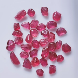 Może przedstawiać: Kolekcja surowych, nieoszlifowanych kamieni szlachetnych rubinu. Kamienie różnią się rozmiarem i kształtem, wykazując gamę odcieni od głębokiego różu do czerwieni. Kamienie są rozrzucone na białej powierzchni, ukazując ich naturalne, szorstkie tekstury i półprzezroczystą jakość.