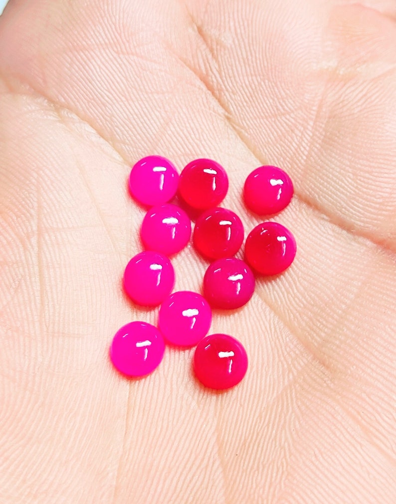 Peut inclure: Une collection de petites perles rondes et brillantes dans des tons de rose et de rouge. Les perles sont dispers&eacute;es sur une surface claire, avec quelques perles d'une teinte rouge plus fonc&eacute;e. Les perles ont un aspect lisse et poli.