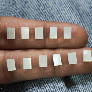 Könnte beinhalten: Zehn kleine, rechteckige Einlegearbeiten aus schillerndem Perlmutt. Jede Einlegearbeit ist etwa 0,6 cm lang und hat einen perlmuttartigen Glanz. Sie sind in zwei Reihen angeordnet. Der Hintergrund ist ein Jeansstoff.