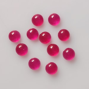 May include: A collection of eleven round, polished, ruby-red gemstones. Each gem has a smooth, reflective surface, with a slight gradient of color from the center to the edges. The gems are scattered on a white surface, showcasing their vibrant color.