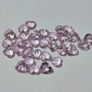 May include: A collection of small, heart-shaped, faceted gemstones in a soft pink hue. The gems are scattered across a white surface, catching the light and displaying multiple facets.
