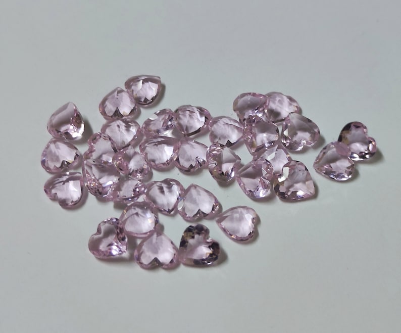 May include: A collection of small, heart-shaped, faceted gemstones in a soft pink hue. The gems are scattered across a white surface, catching the light and displaying multiple facets.