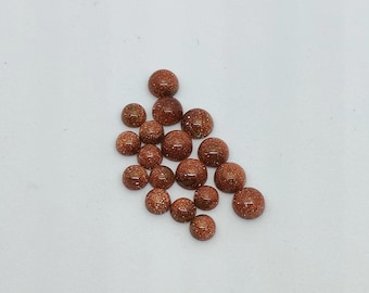 May include: A collection of round, reddish-brown gemstones with a sparkling, glittery appearance. The stones vary in size and are scattered on a white surface. These cabochons could be used for jewelry making or other craft projects.