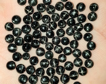 May include: A collection of black star garnet gemstones, each with a prominent four-rayed star pattern. The round stones are polished and reflect light, creating a striking contrast against the background.