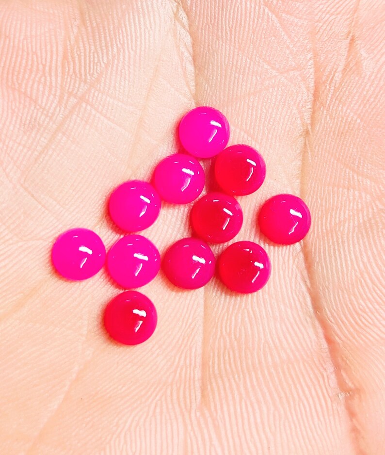 Peut inclure: Un ensemble de cabochons ronds, brillants et roses vifs. Les cabochons sont dispos&eacute;s sur une surface rose clair, mettant en valeur leur aspect lisse et poli. Ces petites gemmes color&eacute;es sont id&eacute;ales pour la cr&eacute;ation de bijoux ou les projets d'artisanat.