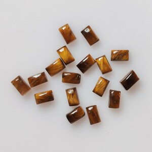 May include: A collection of rectangular tiger's eye gemstones. The stones are a mix of brown and gold, with a polished, glossy finish. The gemstones are scattered on a white surface.