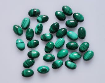 Puede incluir: Colección de gemas de malaquita de forma ovalada. Las piedras pulidas muestran un color verde intenso con patrones arremolinados característicos. Las gemas están dispuestas sobre una superficie clara, mostrando su belleza natural.