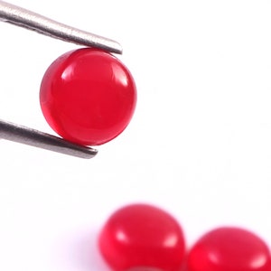 Peut inclure: Gros plan sur une pierre pr&eacute;cieuse ronde rouge vif tenue par une pince argent&eacute;e. La pierre pr&eacute;cieuse est lisse et polie, avec une surface brillante. Deux autres pierres pr&eacute;cieuses rouges sont l&eacute;g&egrave;rement floues en arri&egrave;re-plan, le tout sur fond blanc.