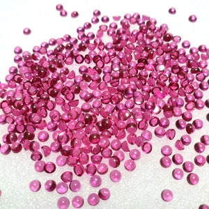 Può includere: Una dispersione di piccole gemme rosa e rotonde. Le gemme sono di un rosa intenso e traslucido, con una superficie lucida e riflettente. Le gemme sono sparse su una superficie bianca.