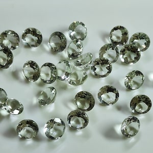 May include: A scattering of oval and round-cut, pale green gemstones on a white surface. The gemstones are translucent, reflecting light and displaying their facets. The arrangement suggests a display of loose gemstones, possibly for jewellery making.