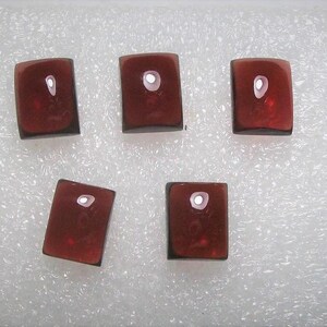 May include: Five rectangular, dark red glass cabochons with a glossy finish and a small, centered, white, oval-shaped inclusion. The cabochons have a dark border and are arranged on a white, textured surface.
