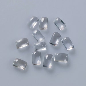 May include: A scattering of clear, rectangular gemstones with rounded edges. The gems are translucent and appear to be made of glass or crystal. They are arranged on a light grey surface, creating a simple, elegant display.