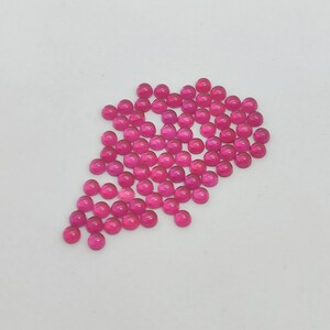 May include: A collection of small, round, cabochon-cut gemstones in a vibrant magenta hue. The gemstones are scattered on a white surface, creating a visually appealing arrangement. These polished stones are suitable for jewelry making or crafting projects.