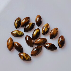 May include: A selection of polished, oval tiger's eye gemstones. The stones exhibit a deep brown hue with golden and yellow chatoyancy, producing a striped, eye-like appearance. The gemstones are displayed on a white surface.