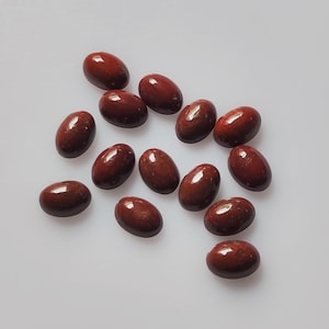 May include: A collection of oval-shaped, polished gemstones in varying shades of reddish-brown. The stones are smooth and reflective, with a glossy finish. They are scattered on a white surface, creating a visually appealing arrangement.