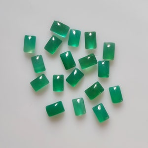May include: A collection of rectangular, polished green gemstones. The stones are evenly cut with rounded edges and a glossy finish. The gemstones are scattered on a white surface, showcasing their vibrant color and clarity. These stones are ideal for jewelry making.