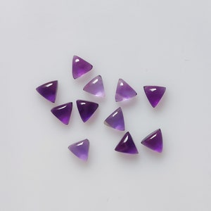 May include: A collection of triangular amethyst gemstones in varying shades of purple. The gemstones are polished and have a smooth, glossy surface. The shapes are uniform, and the stones are scattered on a white background. These gemstones are suitable for jewelry making.