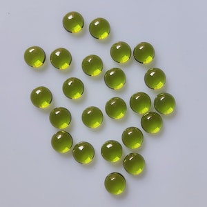 Puede incluir: Una colección de gemas redondas de color verde oliva. Las piedras talladas en cabujón están dispersas sobre una superficie clara. Las gemas tienen un aspecto pulido y brillante, reflejando la luz.
