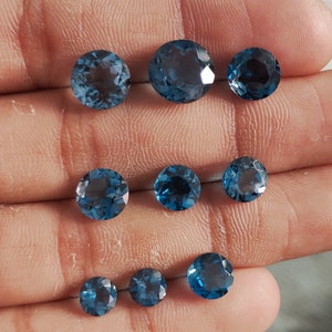 Op de afbeelding: Negen ronde, gefacetteerde, blauwe edelstenen van verschillende grootte worden getoond. De edelstenen hebben een diepe, rijke blauwe kleur en zijn in drie rijen gerangschikt. De edelstenen worden in een hand gehouden, wat hun glans en helderheid laat zien.