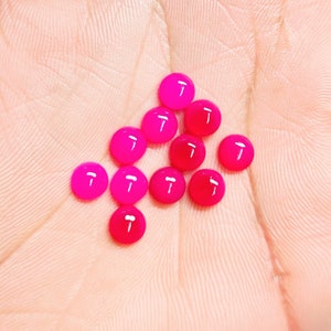 Peut inclure: Plusieurs petits cabochons ronds et brillants, dans des tons de rose et de rouge. Les cabochons sont dispos&eacute;s sur une surface claire, avec une variation de couleur allant du fuchsia vif au cramoisi profond. Les pierres lisses et polies refl&egrave;tent la lumi&egrave;re.