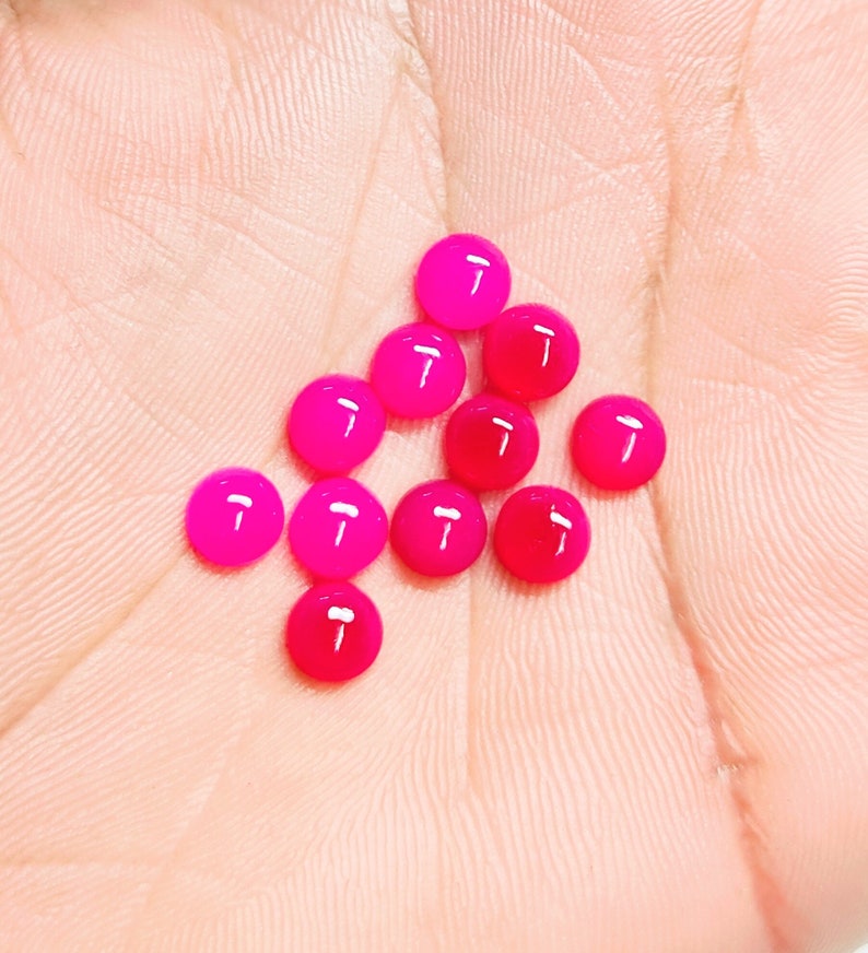 Peut inclure: Plusieurs petits cabochons ronds et brillants, dans des tons de rose et de rouge. Les cabochons sont dispos&eacute;s sur une surface claire, avec une variation de couleur allant du fuchsia vif au cramoisi profond. Les pierres lisses et polies refl&egrave;tent la lumi&egrave;re.