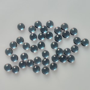 May include: A collection of small, round, translucent, teal-colored glass beads. The beads are scattered across a light-colored surface, reflecting light and creating highlights. These beads could be used for jewelry making or other craft projects.