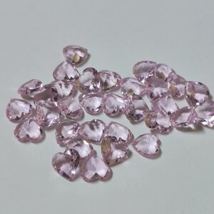 May include: A collection of heart-shaped, faceted gemstones in a soft pink hue. The gemstones are scattered across a light-colored surface, catching the light and displaying multiple facets. The overall effect is one of delicate beauty and sparkle.