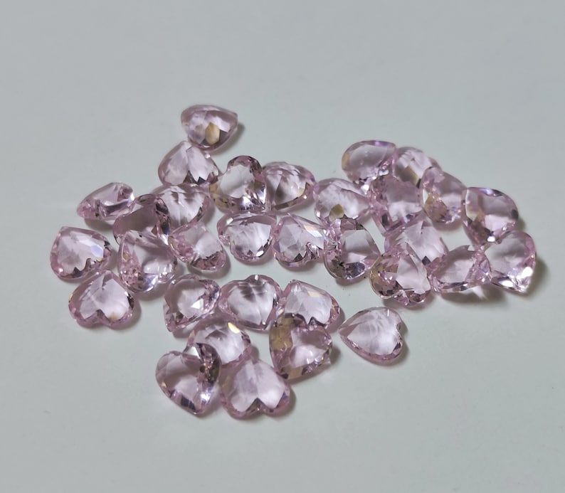 May include: A collection of heart-shaped, faceted gemstones in a soft pink hue. The gemstones are scattered across a light-colored surface, catching the light and displaying multiple facets. The overall effect is one of delicate beauty and sparkle.