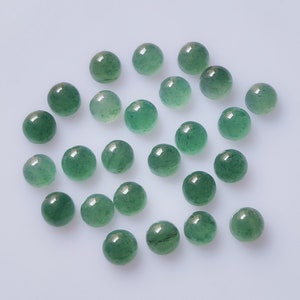 Puede incluir: Una colección de pequeñas piedras preciosas redondas de aventurina verde pulida. Las piedras están dispersas sobre una superficie blanca, cada una con una superficie lisa y reflectante. Las piedras preciosas miden aproximadamente 0,8 cm de diámetro, adecuadas para joyería o manualidades.