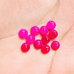 Peut inclure: Une collection de onze pierres pr&eacute;cieuses cabochons rondes, rose vif. Les pierres pr&eacute;cieuses varient l&eacute;g&egrave;rement de teinte, certaines apparaissant plus rouges et d'autres plus roses. Les pierres lisses et polies sont pr&eacute;sent&eacute;es sur un fond de couleur chair.