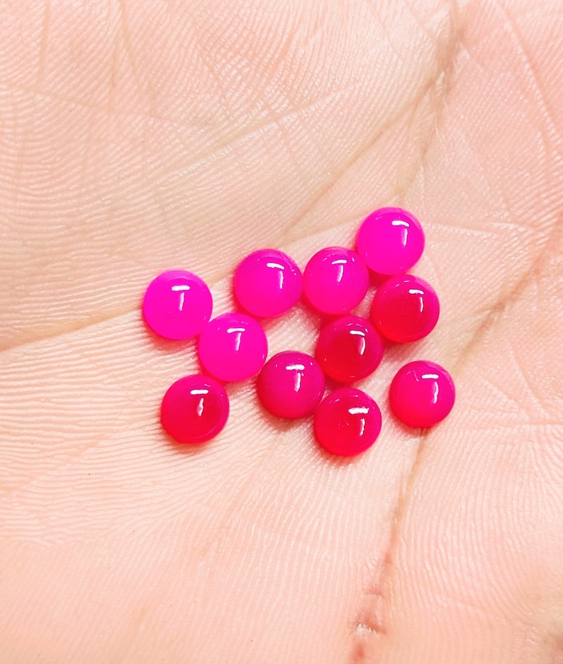 Peut inclure: Une collection de onze pierres pr&eacute;cieuses cabochons rondes, rose vif. Les pierres pr&eacute;cieuses varient l&eacute;g&egrave;rement de teinte, certaines apparaissant plus rouges et d'autres plus roses. Les pierres lisses et polies sont pr&eacute;sent&eacute;es sur un fond de couleur chair.