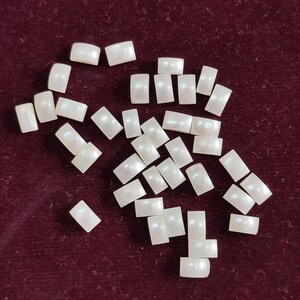 May include: A collection of small, rectangular, iridescent white beads scattered on a dark red surface. Each bead has a slightly curved top and bottom, and a pearlescent sheen.