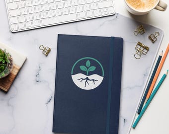 May include: A dark blue notebook with an elastic closure band, featuring a circular graphic on the cover that depicts a green plant sprout with visible roots emerging from white soil. The notebook is positioned on a white marble surface.