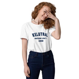 以下が含まれることがあります： ネイビーブルーのブロック体で「KELUTRAL」と書かれた白いTシャツ。その下に「NUMTSENG LENA'I」と書かれています。シャツには「SNL 3744」とプリントされた小さなネイビーブルーの楕円形もあります。モデルは濃い青色のジーンズを着用しています。