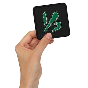 May include: A black square patch with a green and white embroidered design. The design features a stylized 'VS' symbol. The patch is held in a hand, showcasing its size and detail. The edges of the patch are neatly finished.