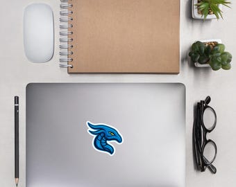 May include: A blue dragon sticker with yellow eyes is affixed to a laptop. The sticker is surrounded by a white border. A white computer mouse, a spiral-bound notebook, a pencil, glasses, and small potted plants are also visible.