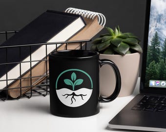 May include: A black ceramic mug featuring a circular graphic on the front. The design consists of a teal-colored sprout with three leaves emerging from a white semi-circle base that displays black, branching roots against a black background.