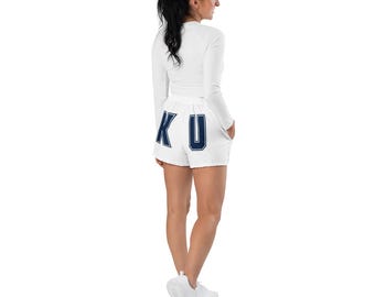 May include: A white athletic outfit featuring a long-sleeved top and shorts. The shorts have the letters "KU" in navy blue on the back. The outfit is worn with white athletic shoes. The person is standing against a white background.