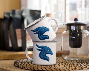 May include: Two white enamel mugs stacked on a woven mat. Each mug features a blue dragon design with yellow eyes. The mugs have silver rims and white handles. A glass teapot and coffee bags are in the background.
