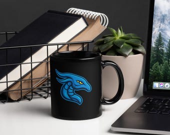 May include: A black ceramic mug featuring a blue dragon design with yellow eyes. The dragon is stylized with a blue outline. The mug is on a white surface next to a laptop and a small potted plant.