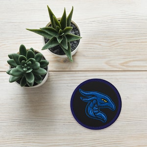 May include: Embroidered patch featuring a blue dragon head on a black background with a navy blue border. The dragon has yellow eyes. Two small potted succulents are also in the image.