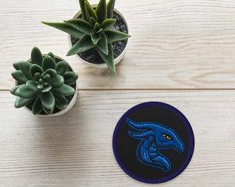 May include: Embroidered patch featuring a blue dragon head on a black background with a navy blue border. The dragon has yellow eyes. Two small potted succulents are also in the image.