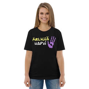 Puede incluir: Camiseta negra con el texto "KELKÜÄ HAPXÌ" en amarillo y un gráfico de mano morada. La camiseta es de cuello redondo y parece estar hecha de un material suave. La modelo lleva vaqueros azules.