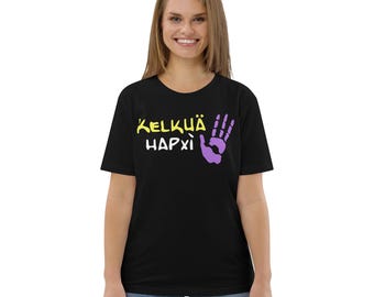 May include: Black t-shirt with the text "KELKÜÄ HAPXÌ" in yellow and a purple hand graphic. The shirt is a crew neck and appears to be made of a soft material. The model is wearing blue jeans.