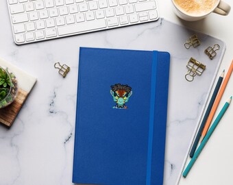 May include: A blue notebook with a vertical elastic closure band features a centered pixel art sticker of a stylized character with dark hair, green skin, and large eyes. The notebook is positioned on a white surface with a keyboard and pencils.