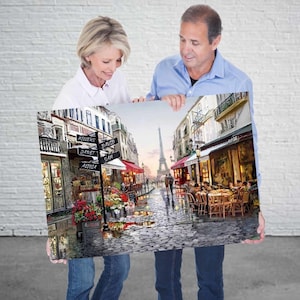 May include: A couple holds up a painting of a Parisian street scene. The painting features a cobblestone street with cafes, shops, and a view of the Eiffel Tower in the distance. The street signs read "Millers", "David Sam", "Janet", and "Aglia".