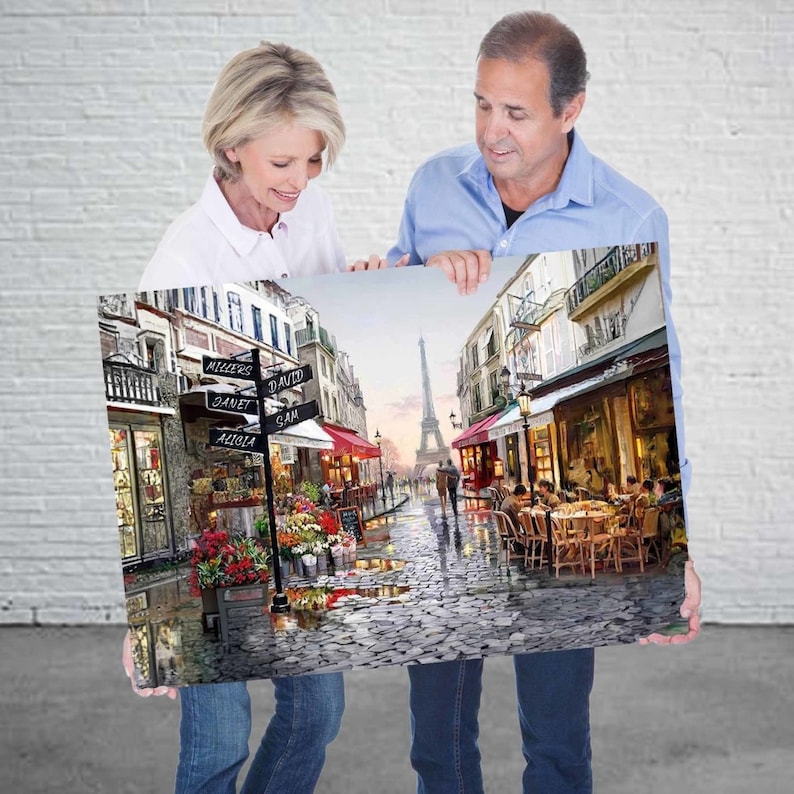 May include: A couple holds up a painting of a Parisian street scene. The painting features a cobblestone street with cafes, shops, and a view of the Eiffel Tower in the distance. The street signs read "Millers", "David Sam", "Janet", and "Aglia".