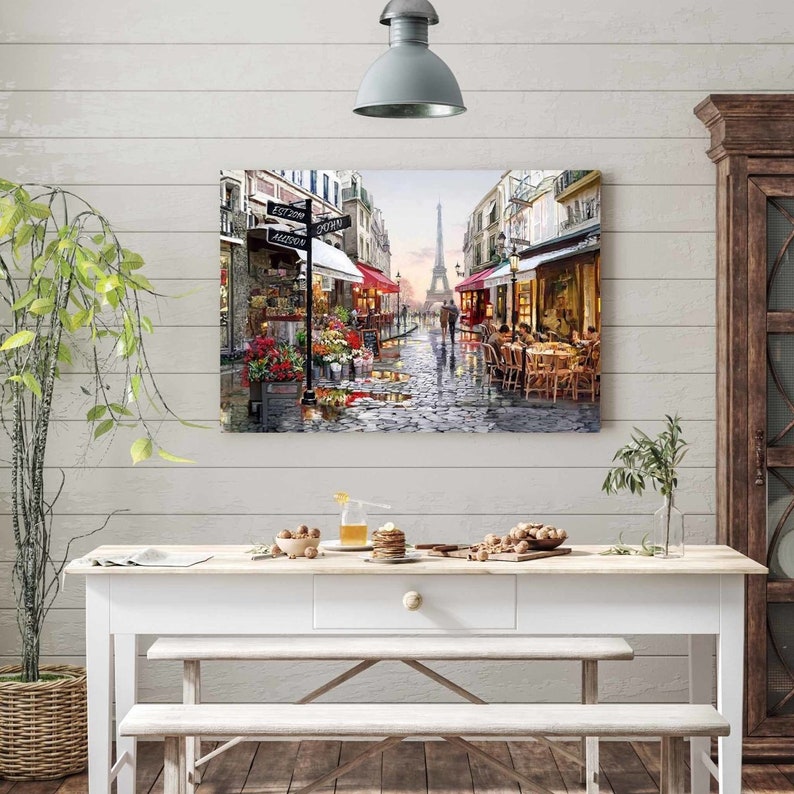 May include: A painting of a Parisian street scene with the Eiffel Tower in the background. The street is cobblestone and there are shops and cafes on either side. People are walking and sitting at tables outside the cafes. The painting is in a realistic style and has a warm, inviting feel.