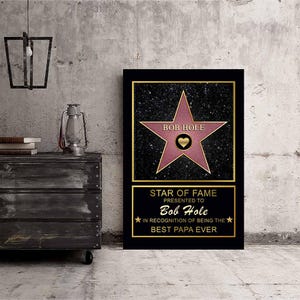 Op de afbeelding: Een zwart en gouden ingelijst afdruk met een roze ster op een zwarte achtergrond. De ster heeft de tekst "BOB HOLE" en een hart in het midden. De tekst onder de ster luidt "STAR OF FAME PRESENTED TO Bob Hole IN RECOGNITION OF BEING THE BEST PAPA EVER".