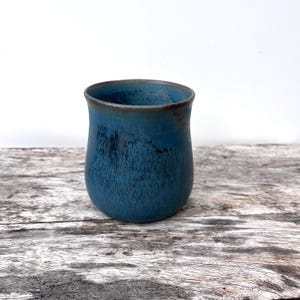 Könnte beinhalten: Ein Keramikbecher mit einer abgerundeten, bauchigen Form und einer weiten Öffnung. Die Außenseite ist mit einer tiefblauen Glasur überzogen, die dunklere, texturierte vertikale Streifen und einen dezenten bräunlichen Rand aufweist.
